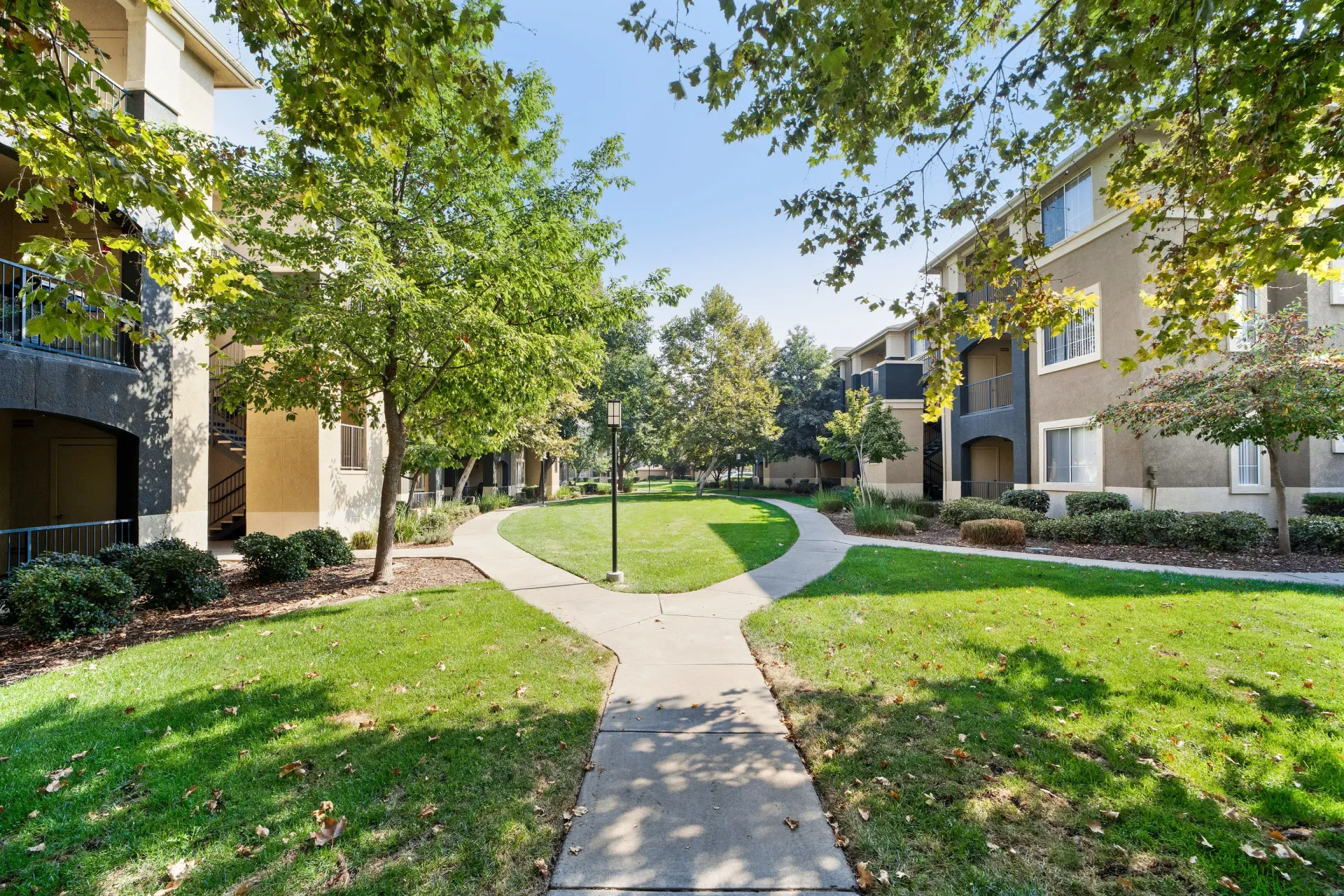 Outdoor community courtyard with trees, green lawn, and curved walkways between apartment buildings.