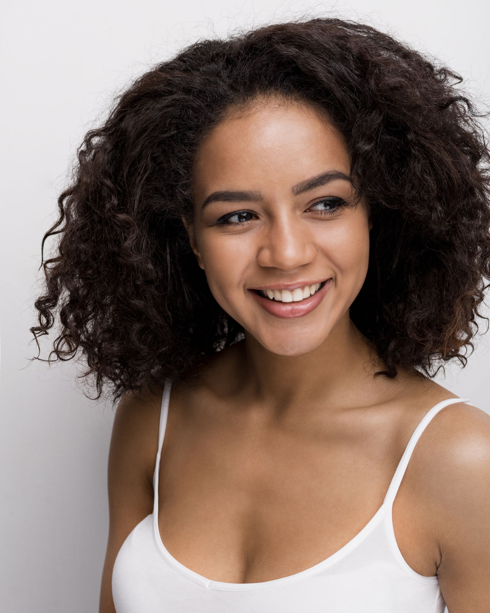 a woman with curly hair is smiling and wearing a white tank top .