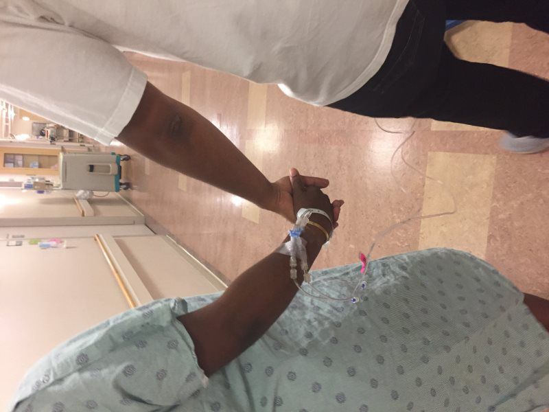 Person in hospital bed, hand held by another. IV line visible.