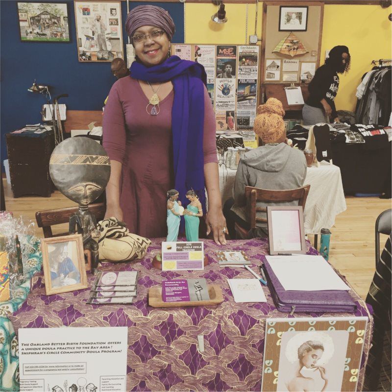 Woman at a table with items for sale; purple and blue accents, smiling. Indoor setting.