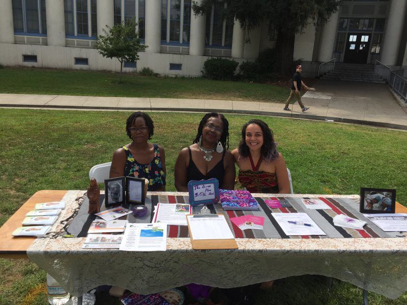 Three women seated at a table outdoors, smiling. Table has items displayed on a decorated cloth, building in background.