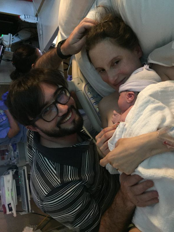 Parents smiling, holding newborn baby wrapped in a blanket in a hospital setting.