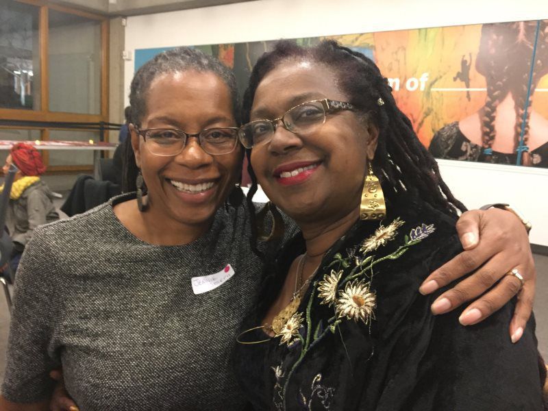 Two women smiling, one with arm around the other. Indoors, artwork in the background.