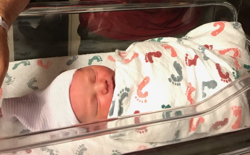 Newborn baby in hospital bassinet, wrapped in blanket with red and green footprints.