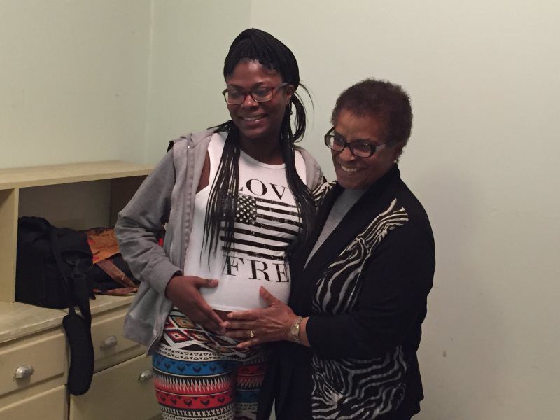 Pregnant person with a companion touching belly. They are smiling inside a room with a light-colored wall.