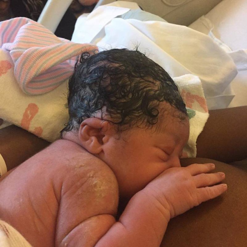 Newborn baby resting on someone's chest with dark, wet hair; hospital setting.