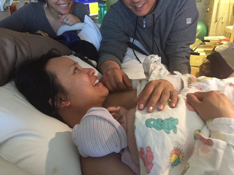 Woman in bed laughing, holding baby, surrounded by smiling people.