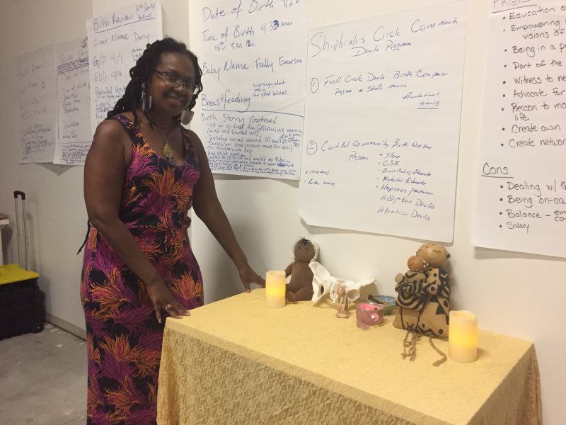 Woman at a table with candles and figures; whiteboards behind her.
