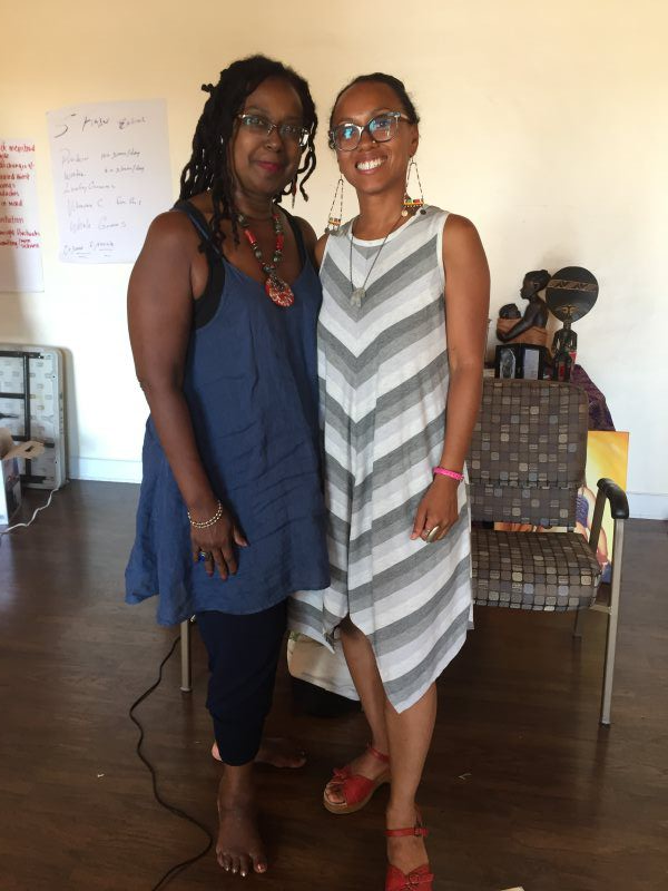 Two people smiling, posing together indoors. Woman on the left wears blue dress; woman on the right wears striped dress.