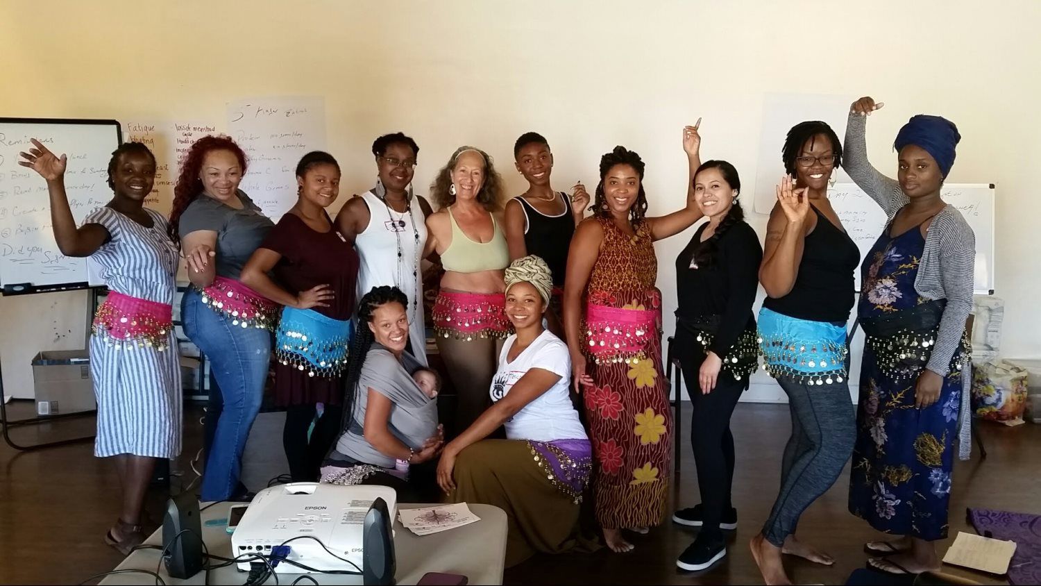 Group of people in a room, some wearing colorful hip scarves, posing and smiling.