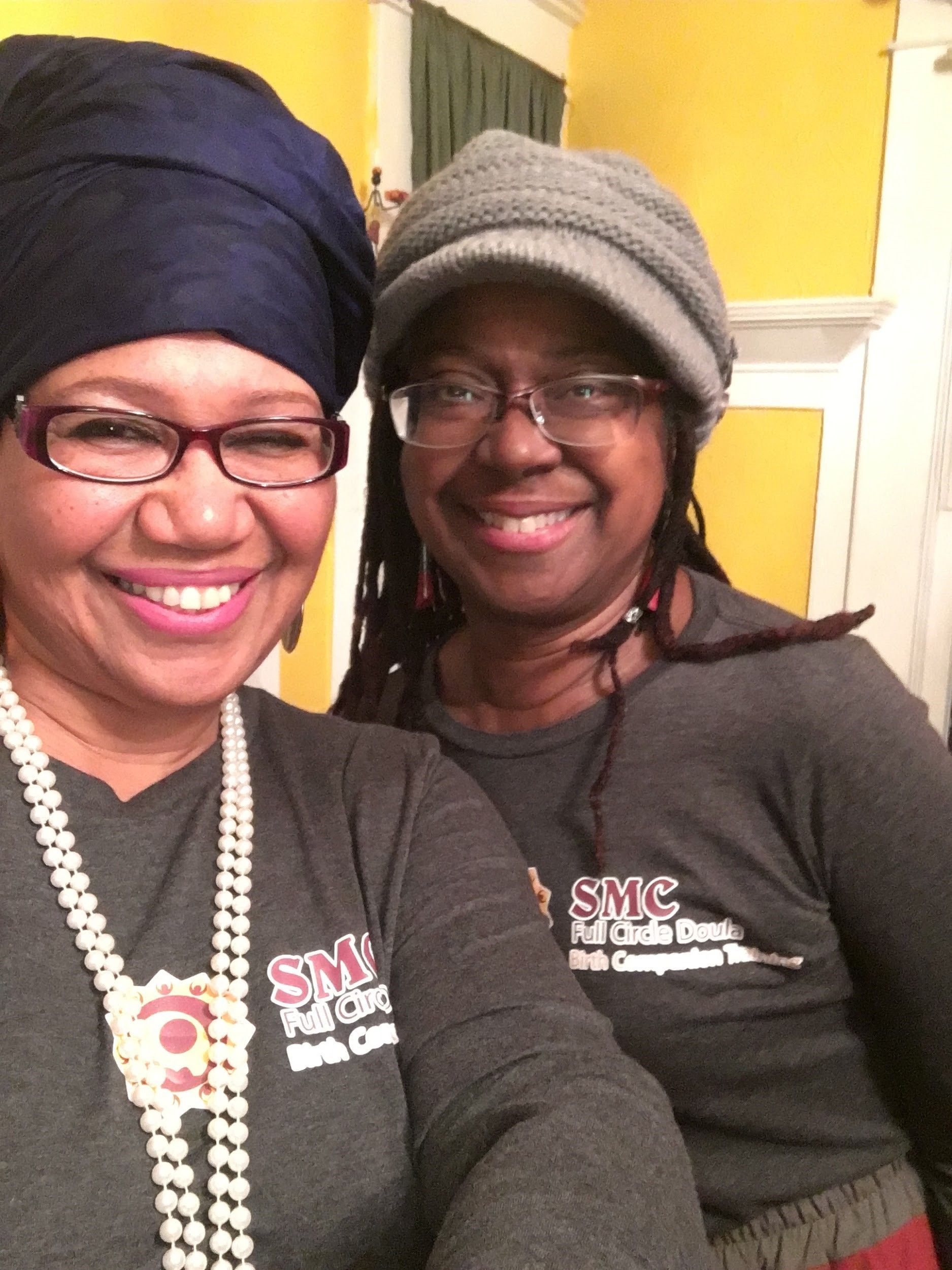Two smiling people in a close-up photo. One wears a blue headwrap and glasses. The other has dreadlocks and a gray cap.