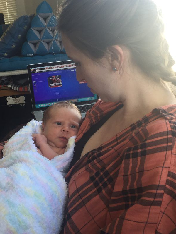 Woman holding a swaddled baby, looking at a laptop screen in a room with natural light.