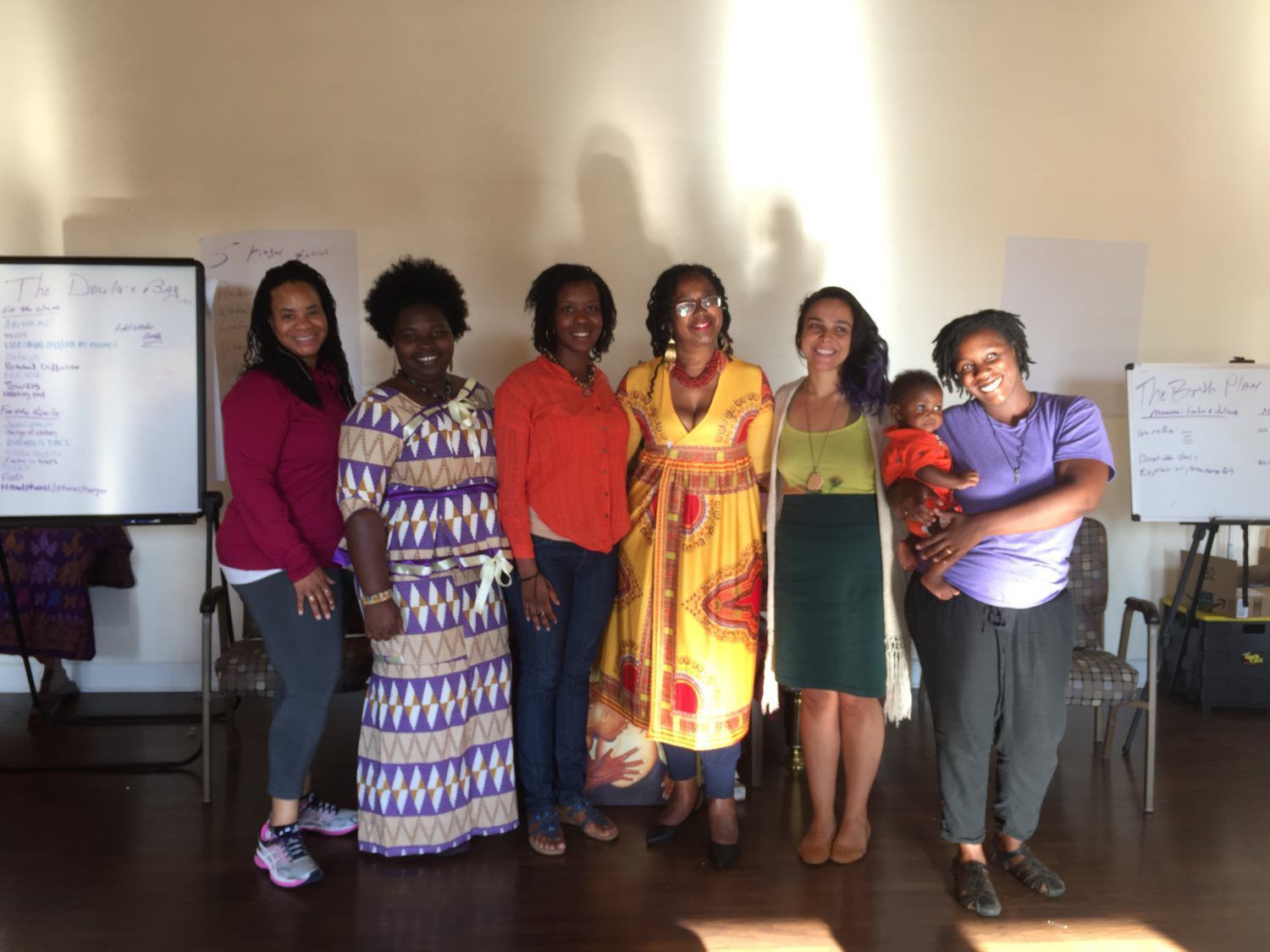 Group of women stand together in a room with whiteboards. They are smiling. One woman holds a baby.