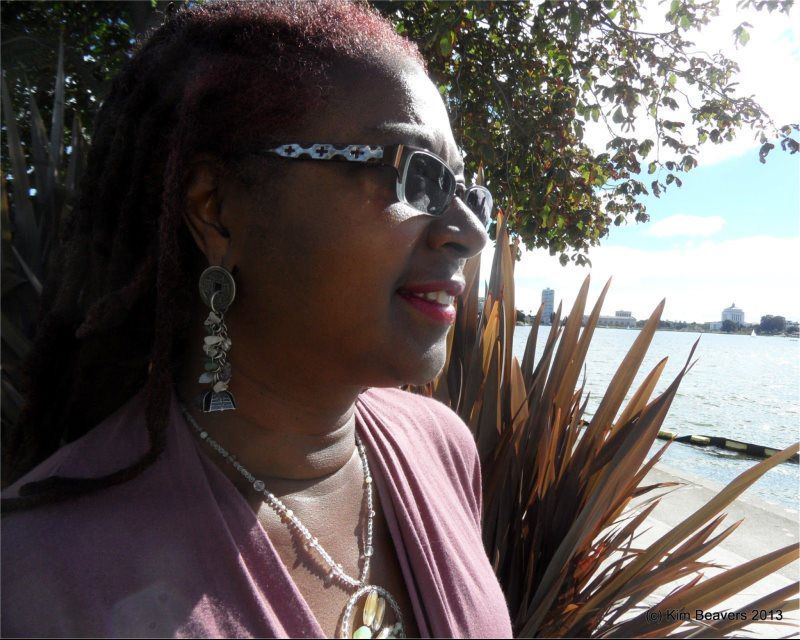 Woman in sunglasses looks towards water. Pink shirt, silver jewelry, and red lipstick. Sunny day.