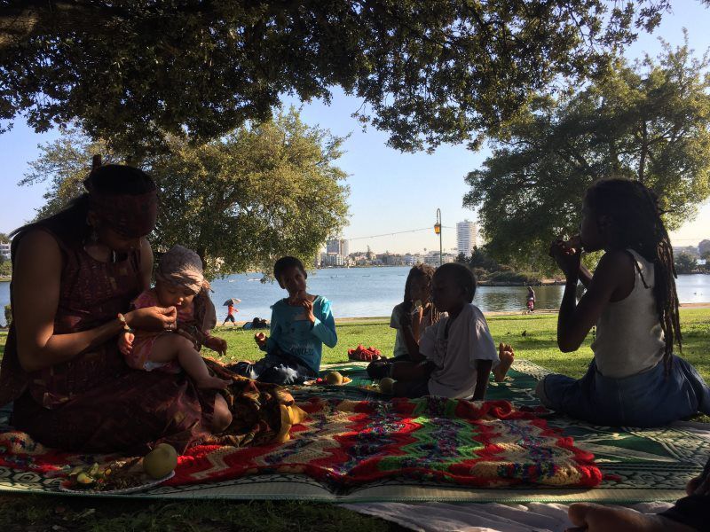 People sitting on a blanket, eating by a lake under trees. Bright sunny day.