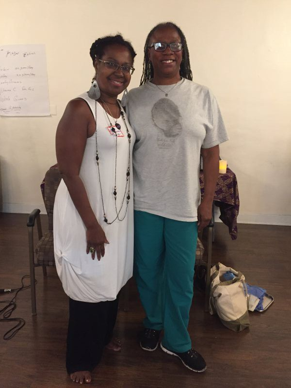 Two people smiling, standing indoors. One in white dress, black pants; the other in gray shirt, teal pants.