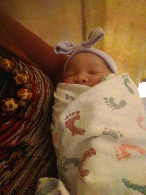Newborn baby wrapped in a blanket with footprints, wearing a purple hat, held by a person.