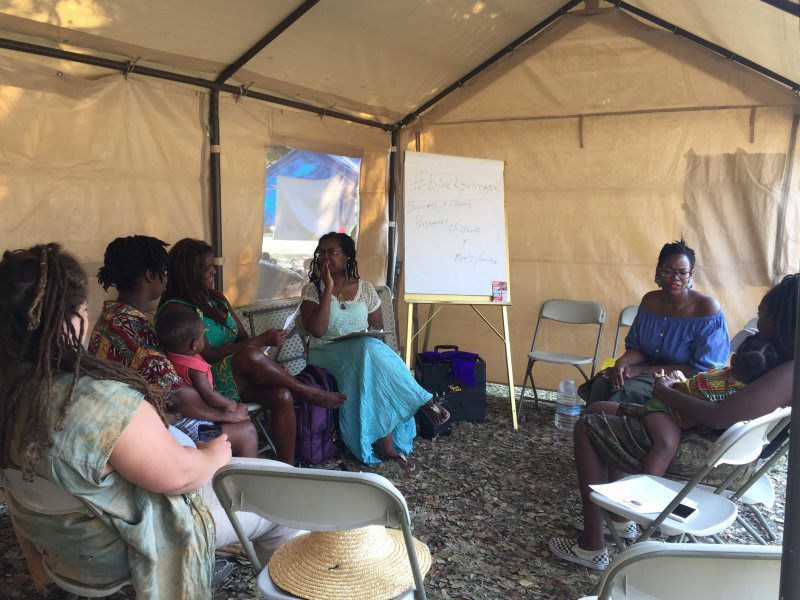 Group of women in a tent having a discussion; woman with whiteboard is speaking.