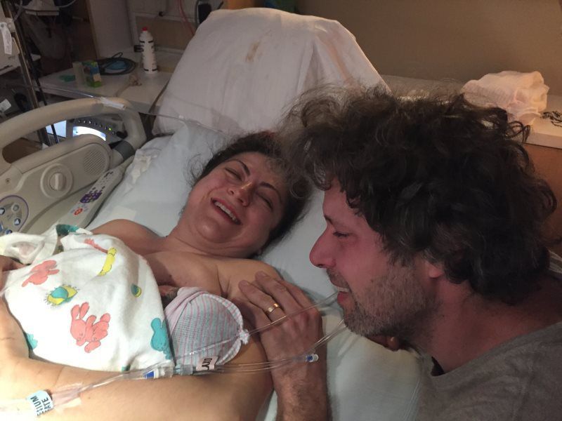 Woman in hospital bed smiles, holding newborn; man beside her smiles, touching her hand.