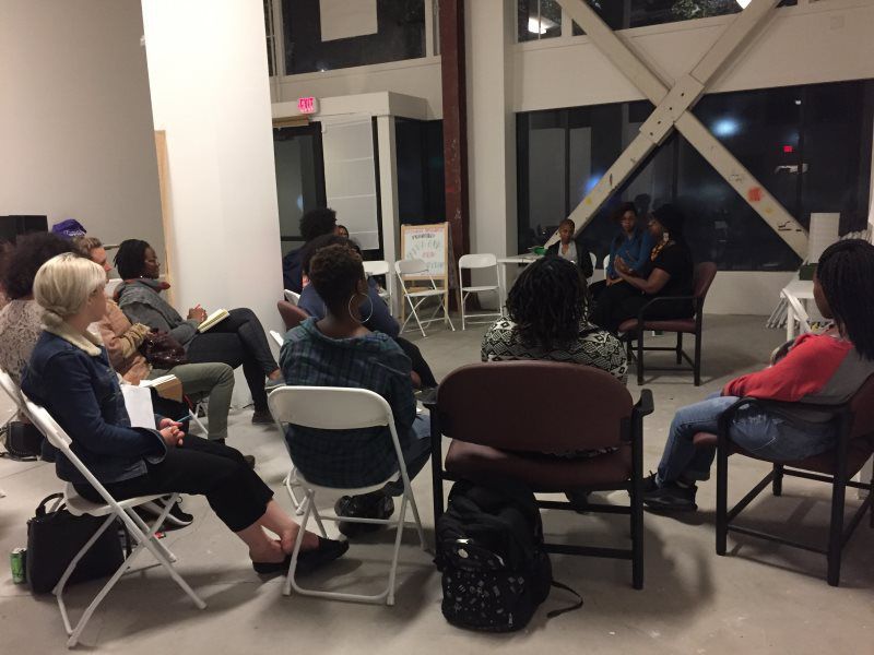 People seated, listening to two women. A discussion group is taking place in a room with a concrete floor.