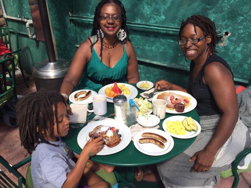 Three people eating breakfast at a green table outdoors. Plates of food and drinks are visible.