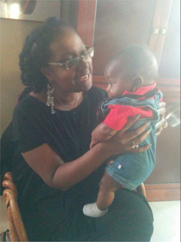 Woman holding a baby, both looking at each other. The woman wears glasses and earrings. Baby in a red shirt and overalls.