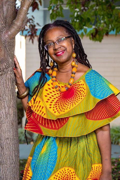 Woman wearing vibrant African print dress, smiles while leaning on a tree.