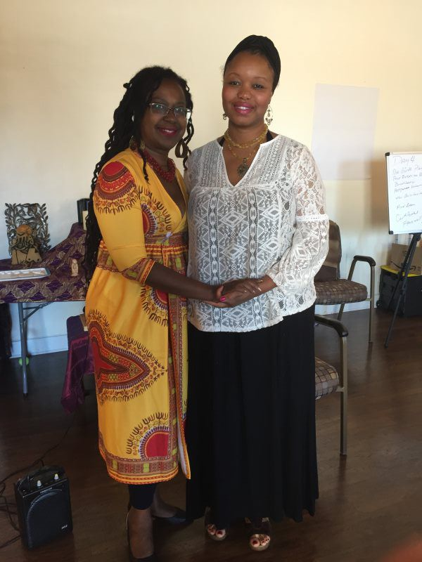 Two women holding hands, one in yellow patterned dress, the other in white lace top and black skirt, inside.