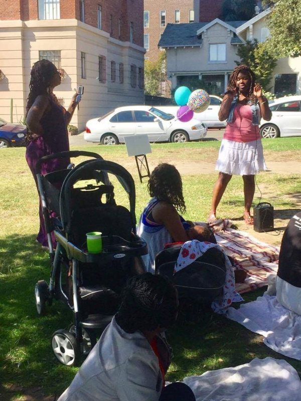 People at a park picnic: one woman speaks, others seated, baby stroller, balloons. Cars and buildings in the background.