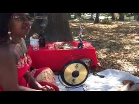 Woman sitting outdoors near red table with gong and ritual objects.