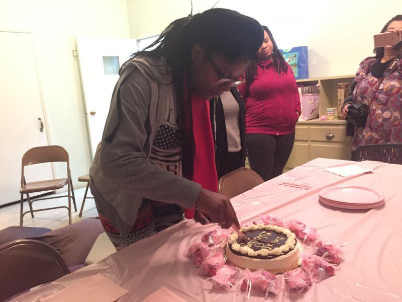 Person cutting a cake at a party. Pink tablecloth, guests watching.