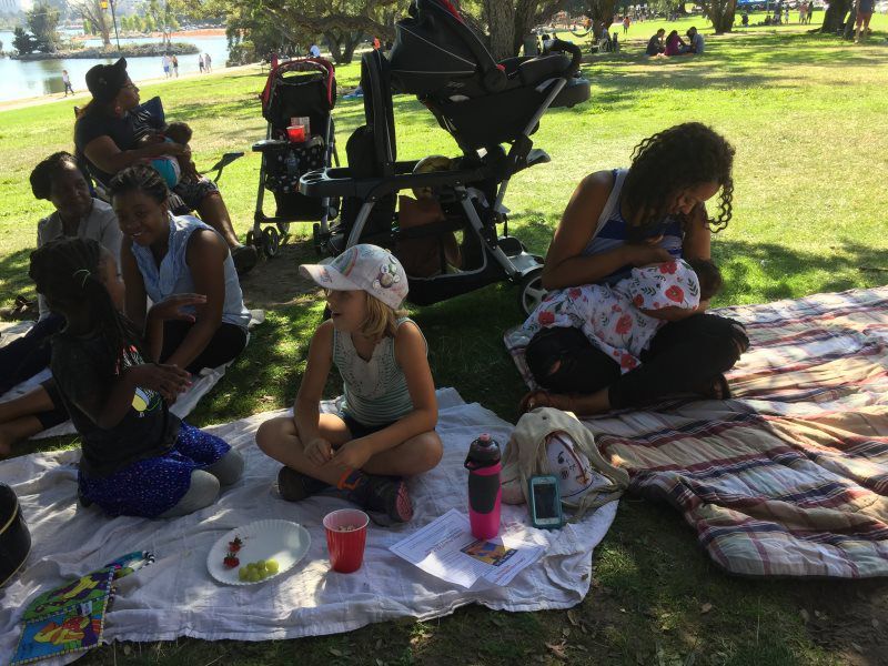 People picnicking in a park; a woman breastfeeding a baby, others sit on blankets, strollers nearby, sunny day.