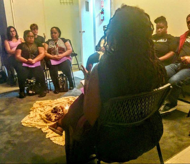 Group of people in chairs, one in the foreground facing the others with dreadlocks, ritual items on a cloth in the center.