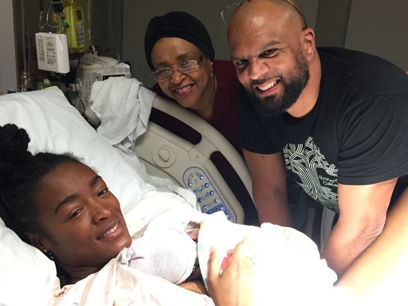 Woman in hospital bed smiles, with smiling man and older woman leaning in, holding baby.