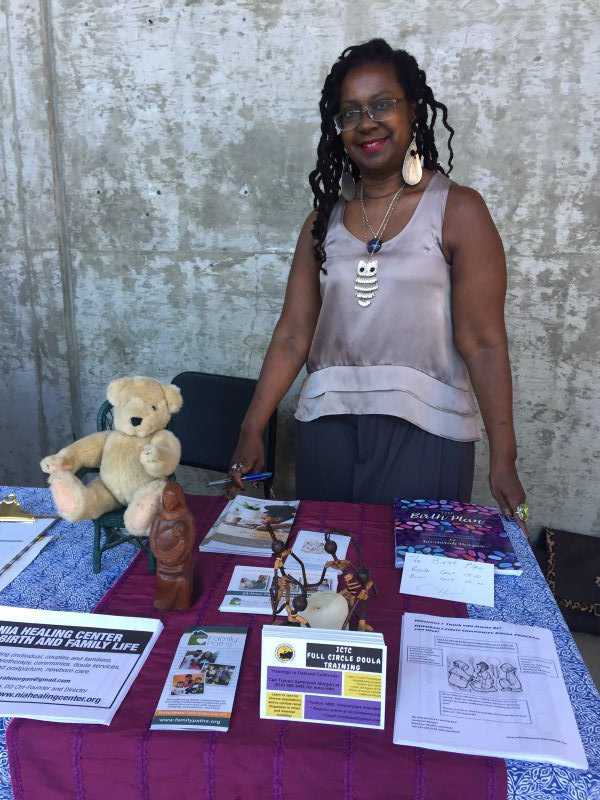 Woman standing at a table with brochures, figurines, and a teddy bear. She smiles, leaning on the table outdoors.