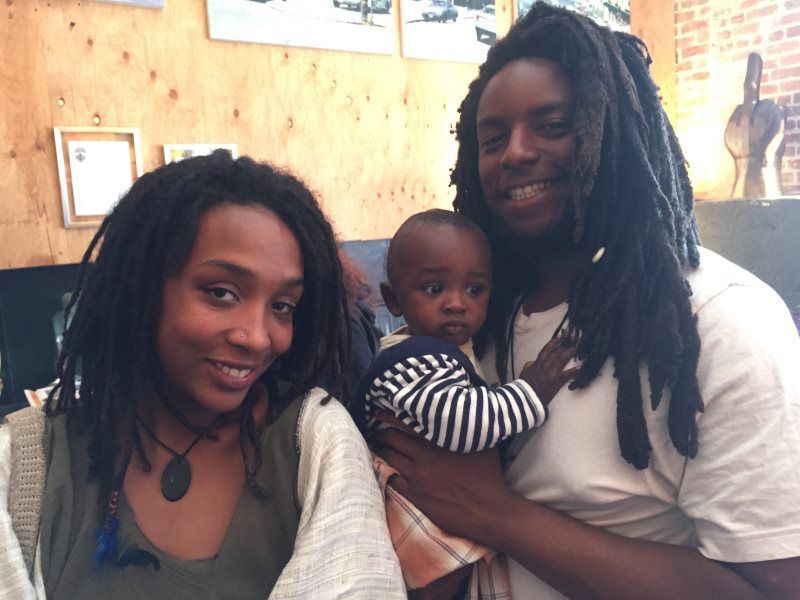 Family with dreadlocks smiling, holding a baby, seated in front of a wood-paneled wall.
