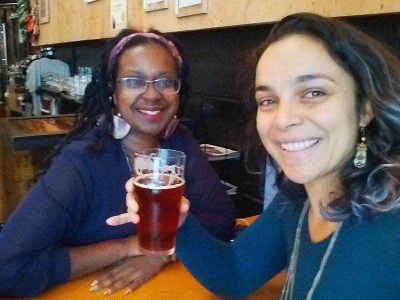 Two women smiling, toasting beer glasses in a bar with wooden accents.