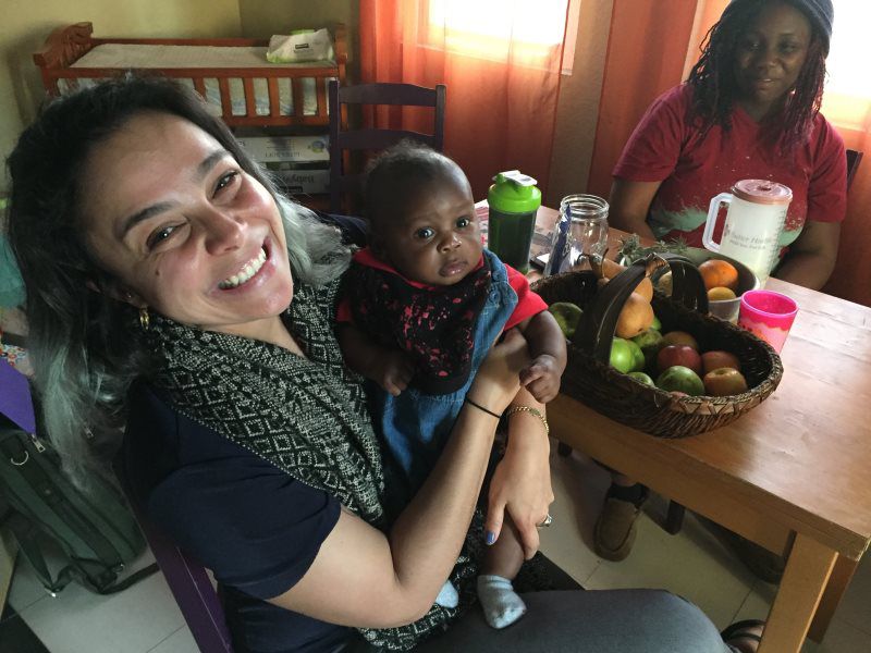 Woman holding a baby, smiling. Another woman sits at a table with fruit. Interior setting.
