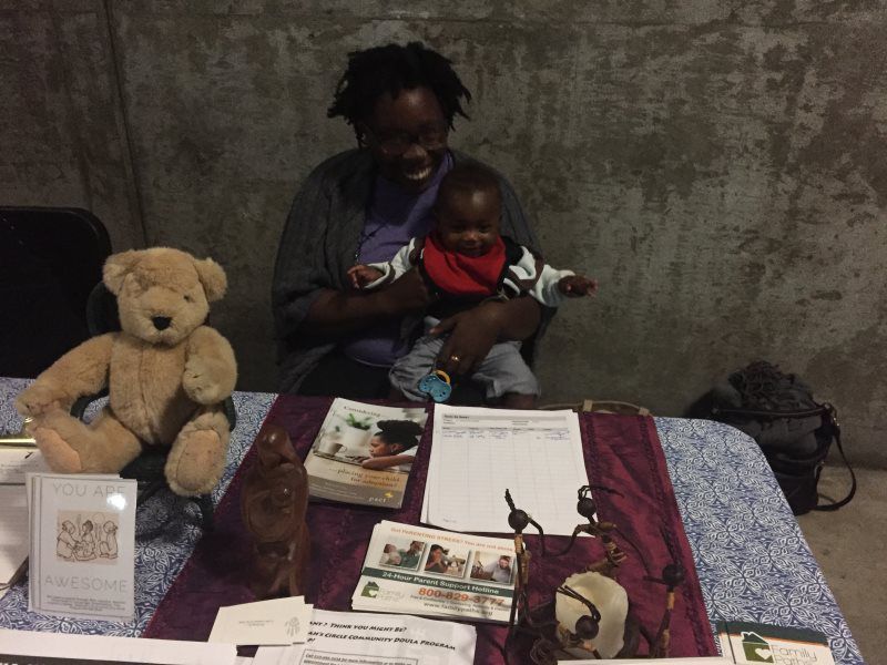 Woman holds baby at a table display with books, teddy bear, and artwork against a concrete wall.