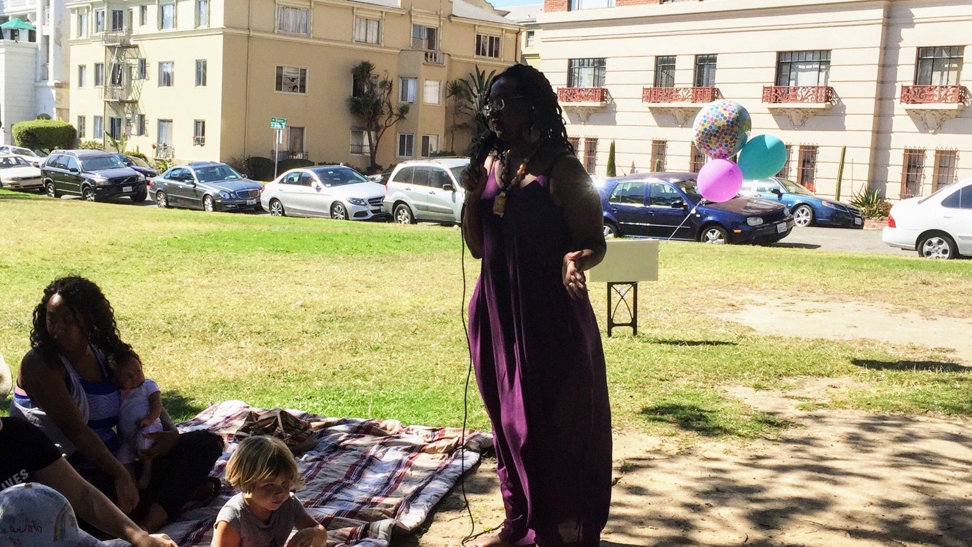 Woman in purple dress speaking into a microphone in a park. People sit on a blanket; cars line the street.