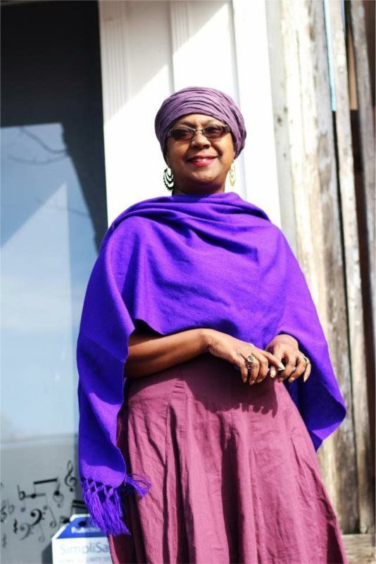 Woman in purple headscarf and shawl, standing outside.