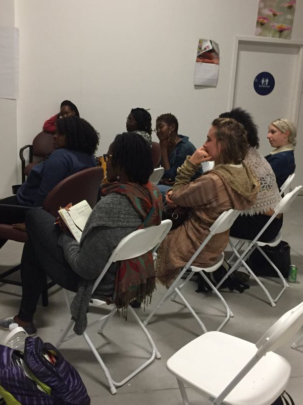 People seated in folding chairs, gathered indoors, some looking forward, a few with notebooks.