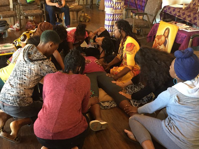 A group kneels around a person lying down, hands on body. Indoor setting, various colors.