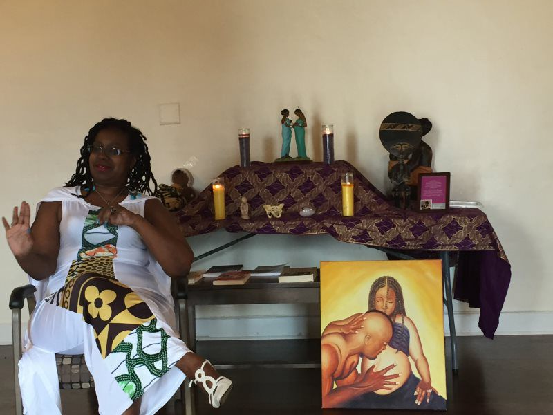 Woman sitting, gesturing with hand, near a table with candles and an art piece depicting two figures.