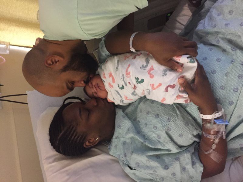Parents and newborn in hospital bed, touching noses. Baby is swaddled in a patterned blanket.