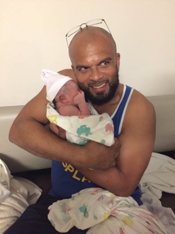 Man holding a newborn baby wrapped in a blanket. The man smiles and wears glasses on his head.