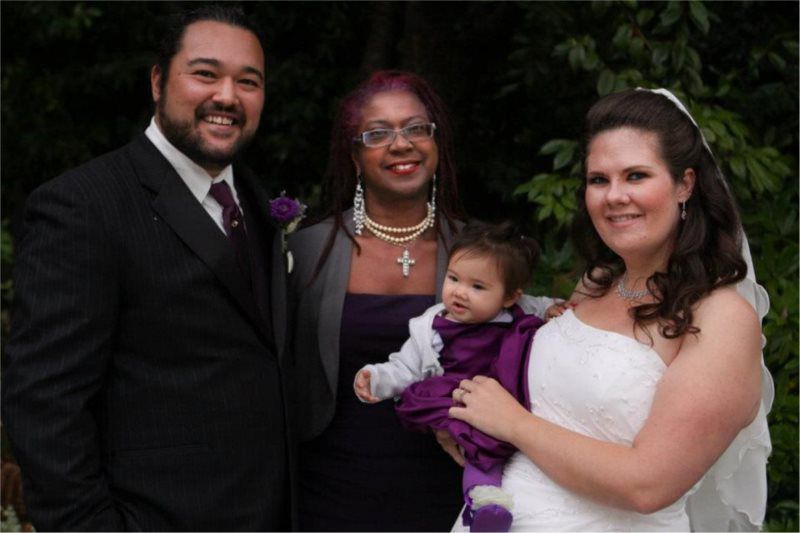 Wedding group: Man in suit, bride in white dress, woman holding baby, woman in purple, all smiling outdoors.