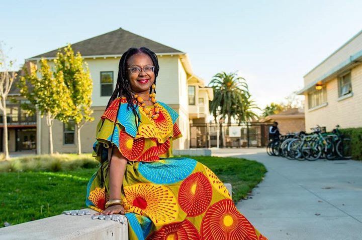 Woman in colorful dress sits outdoors, smiling. Building, walkway, and bikes are in background.