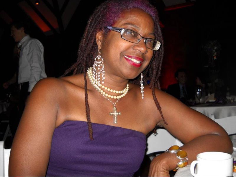 Woman with purple dreadlocks wearing glasses, purple dress, and jewelry smiling at a formal event.