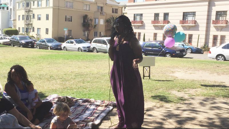 Woman in purple dress speaking into a microphone in a park. People sit on a blanket; cars parked nearby.